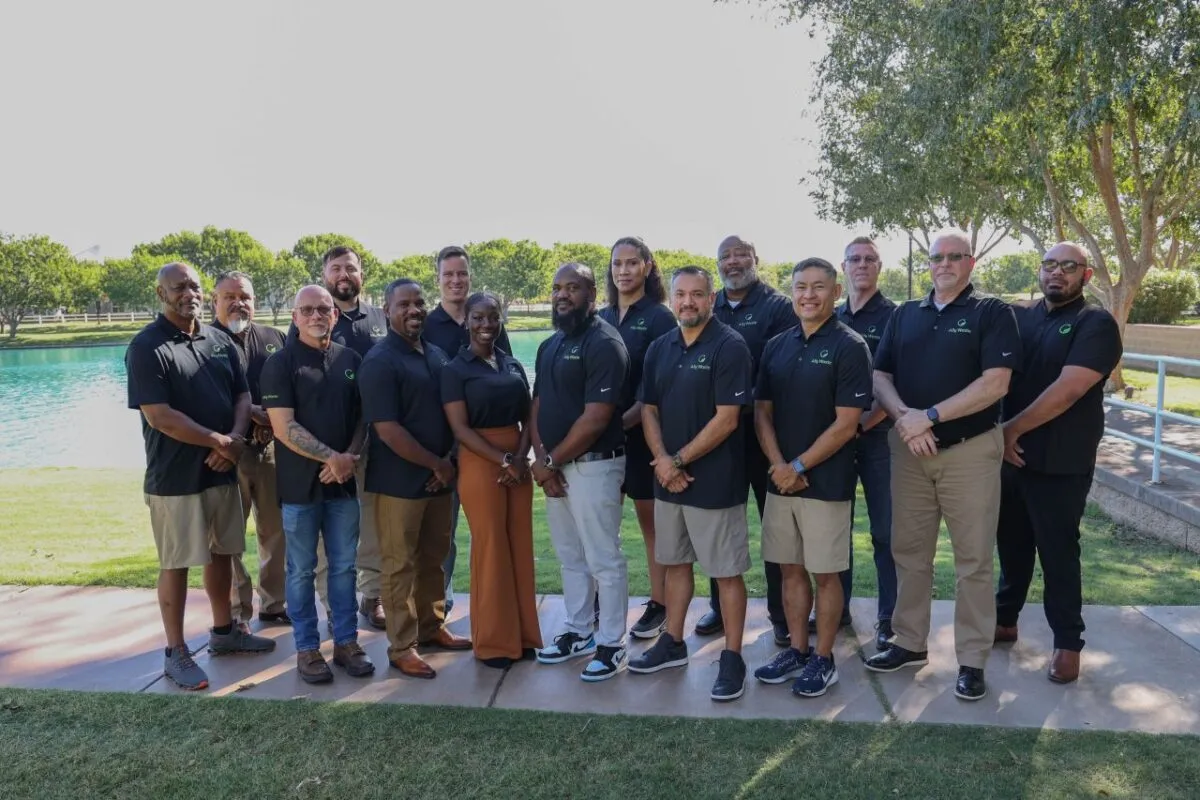 About twenty Ally Waste team members in matching black polos, standing together on a lawn by a lake